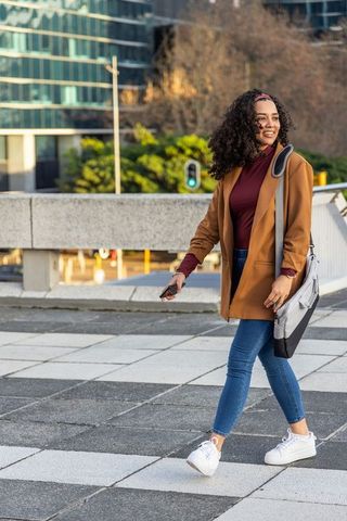 Young woman walking urban plaza in camel coat carrying shoulder bag and checking phone