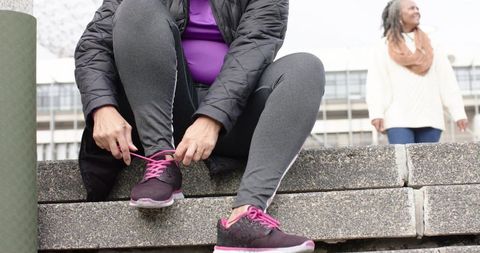 Mature woman tying pink shoelaces on purple sneakers on urban steps with friend watching