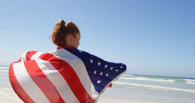 Young woman standing at sandy beach with American flag draped over her shoulders. Ideal for themes related to patriotism, summer vacations, beach holidays, freedom, and national celebrations in the United States.