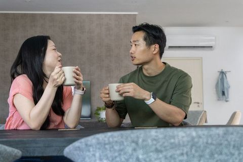 Asian couple talking and sipping coffee at dining table in cozy minimalist apartment
