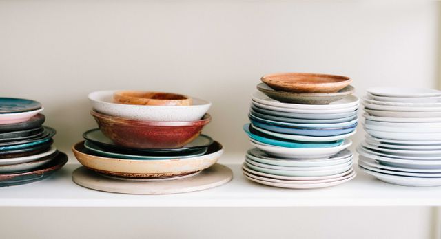 Assorted ceramic dishes, including plates and bowls, are neatly stacked on white shelves. This assortment showcases a variety of colors and textures, creating a visually appealing arrangement. Perfect for use in articles or advertisements about kitchen organization, home decor, or culinary topics, highlighting tidiness and aesthetics in the kitchen environment.
