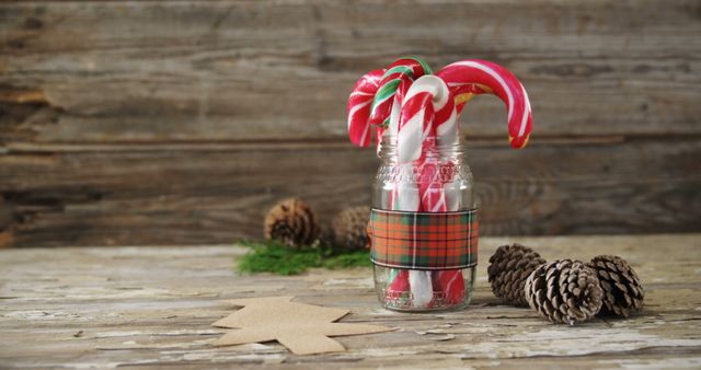 Candy canes are nestled in a jar with a festive plaid pattern, evoking the warmth of holiday traditions, with copy space. Pinecones and a rustic wooden backdrop add to the cozy, seasonal vibe.