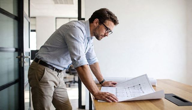 Architect reviewing blueprints on minimalist office desk, wearing blue shirt studying plans