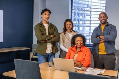 Diverse team collaborating around laptop in modern office meeting room, smiling leaders