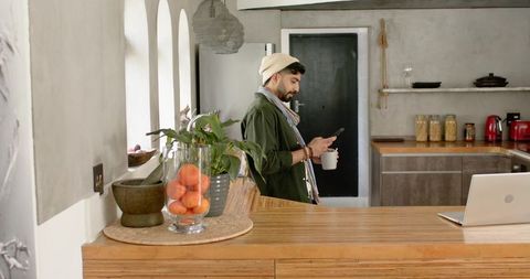 Indian man relaxing in modern rustic kitchen holding coffee and checking smartphone