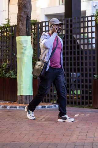 African american man walking brick sidewalk talking on smartphone wearing cap and crossbody bag