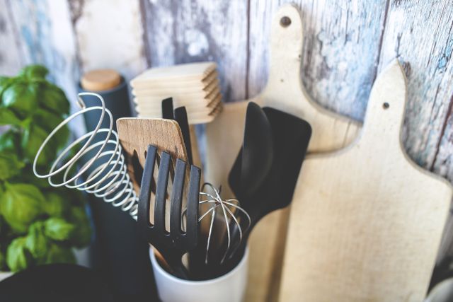 Various kitchen utensils like spatulas, whisks, and ladles are stored in a container, set against a wooden background with cutting boards nearby. Perfect for articles or blogs on home cooking, kitchen organization, rustic kitchen decor, or culinary tools.