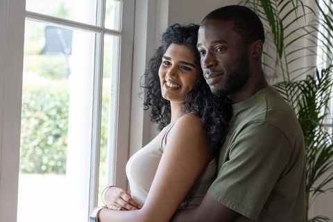 Indian woman and african american man embracing and smiling by sunlit window, cozy couple