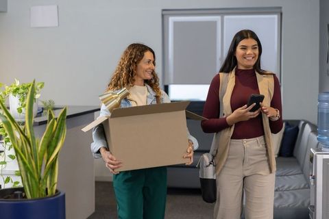 Diverse female coworkers walking through office reception carrying box and brass lamp