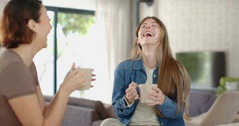 Laughing friends sharing coffee in cozy living room holding mugs casual conversation