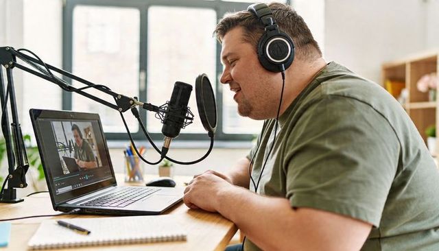 Male podcaster recording from home studio on laptop with microphone and headphones