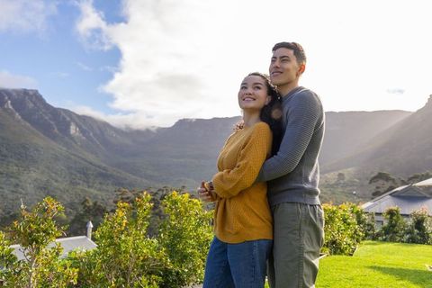 Asian couple embracing on sunlit mountain lawn overlooking valley and peaks