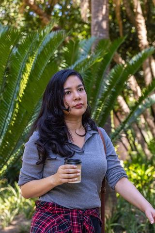 Woman walking through tropical garden holding takeaway coffee cup in casual outfit