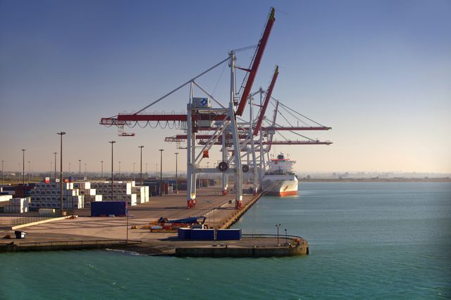 Large seaport terminal showing shipping containers and massive crane system for loading and unloading cargo ships. Ideal for illustrating concepts related to global trade, logistics, international commerce, and marine transportation. Could be used in articles about supply chain, industrial operations, or economic trade reports.