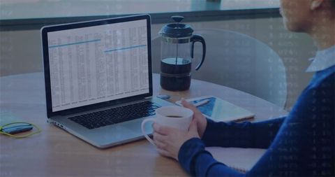 Woman sipping coffee while reviewing spreadsheet on laptop in modern bright workspace