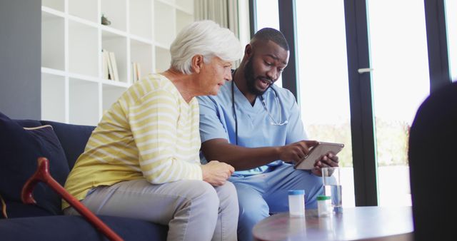 Image of african american male doctor using tablet with caucasian senior woman. seniors health and nursing home lifestyle concept.
