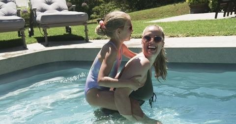 Mother and daughter playing in backyard pool wearing one-piece swimsuits and sunglasses
