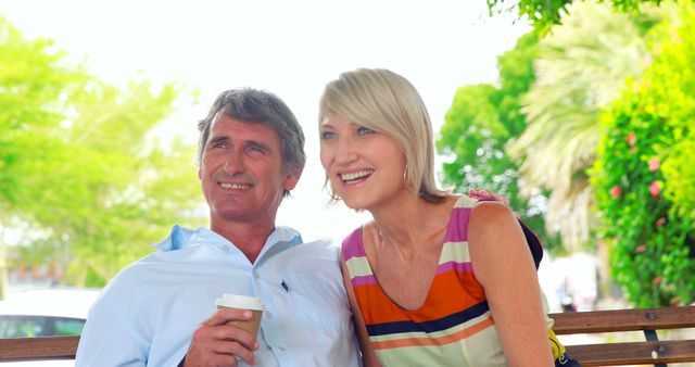 Senior couple sitting on a park bench, enjoying coffee and each other's company. Both are smiling and appear relaxed. Can be used in contexts related to retirement, leisure activities for seniors, happy relationships, and spending quality time outdoors.