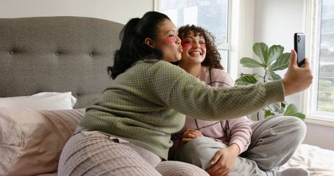 Friends taking selfie on bed with under-eye patches together enjoying cozy skincare moment