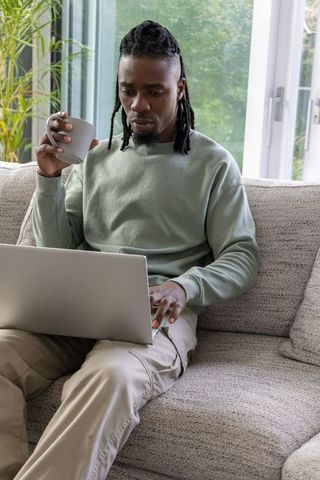 African american man working from home on laptop holding coffee mug on sofa minimalist
