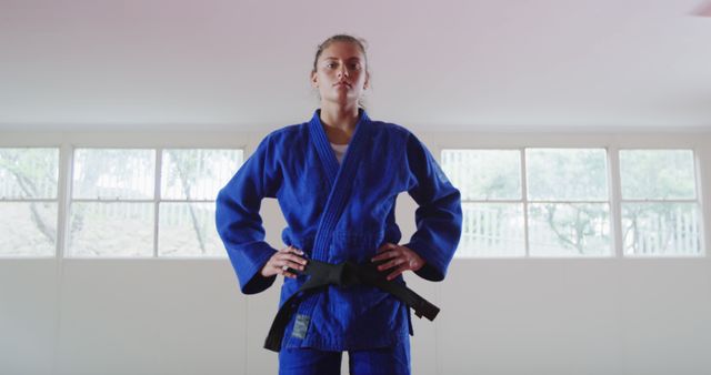 Confident woman wearing blue gi and black belt, standing with hands on hips in dojo. Ideal for themes related to martial arts, women's empowerment, fitness training, self-defense, discipline, and strength. Useful for promotion of martial arts classes, fitness programs, and women's self-defense courses.