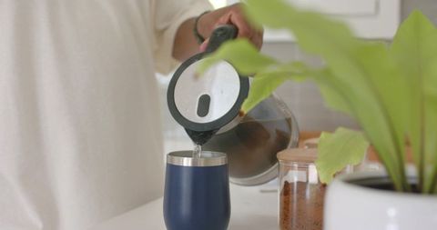 African man pouring brewed coffee into navy travel tumbler on minimalist kitchen counter at home