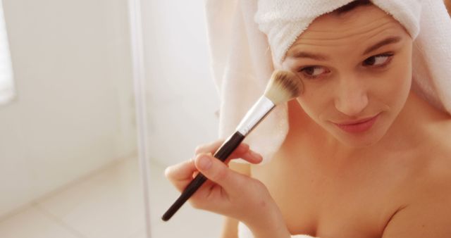 This image shows a woman in a bathroom applying makeup with a brush after a shower. She has a towel wrapped around her head, indicating a fresh and clean feeling. Ideal for use in beauty tutorials, skincare advertisements, cosmetics marketing, and health and wellness articles.