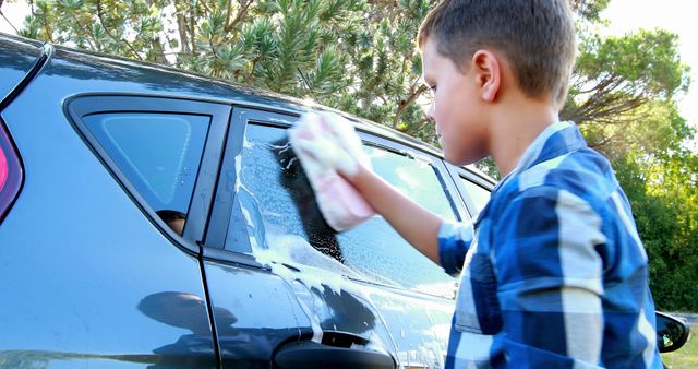 A young Caucasian boy is washing a car window with a sponge, working diligently in a sunny outdoor setting. His involvement in the chore demonstrates responsibility and the importance of contributing to household tasks from an early age.