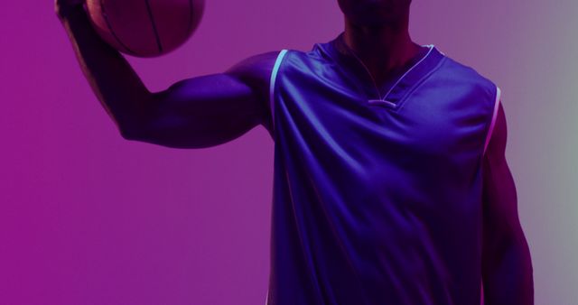 Athlete holding a basketball in vibrant neon purple light, showcasing a muscular arm and wearing a sports jersey. This image is ideal for use in fitness, sports promotions, athletic event posters, or marketing materials highlighting sportswear brands.