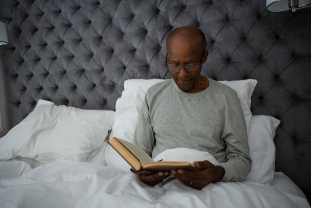 Senior man sitting in bed reading a book, creating a peaceful and cozy atmosphere. Ideal for use in articles or advertisements about senior living, relaxation, leisure activities, or promoting reading habits among elderly individuals. Can also be used in healthcare or lifestyle blogs focusing on the well-being of mature adults.