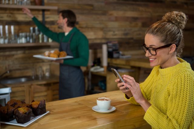 Happy woman using mobile phone while barista working in background at coffee shop