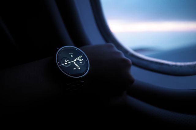 Close-up of a wristwatch worn by a person resting near an airplane window during dusk. Suitable for use in travel-related content, luxury lifestyle promotions, time management concepts, and adverts highlighting stylish accessories or technology in everyday life.