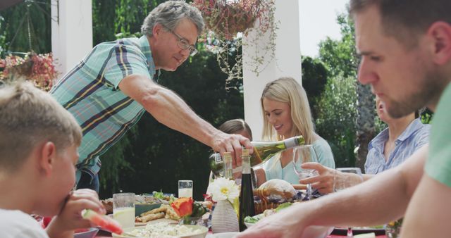 Happy caucasian family sitting at table in garden, eating dinner and drinking wine. Lifestyle, domestic life, family, and togetherness.