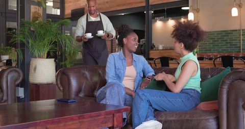 African American Friends Sitting on Sofa Talking in Cozy Cafe While Barista Bringing Coffee