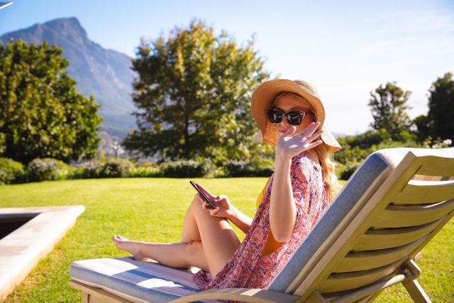 Caucasian woman sitting on a sunbed in a garden, wearing sunglasses and a hat, using a smartphone. Perfect for lifestyle blogs, vacation advertisements, technology in leisure contexts, and summer relaxation themes.