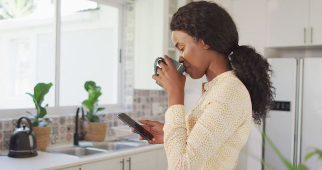 Young African American woman stands in a modern kitchen, drinking coffee while using her smartphone. Sunlight filters through the window, creating a bright, clean atmosphere. Perfect for depicting morning routines, modern lifestyle, home and tech usage, or casual indoor settings.