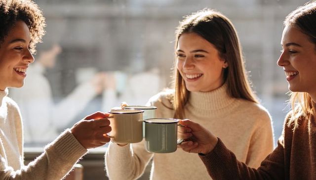 Friends clinking enamel mugs in sunlit cafe wearing cozy knit sweaters, sharing laughter