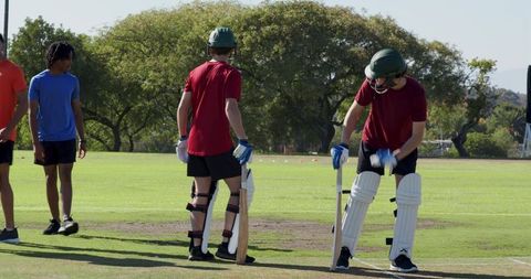 Young cricketers adjusting pads and gripping bats on grassy practice pitch with teammates