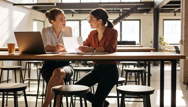 Two women collaborating over documents in sunlit open-plan office with laptop and coffee