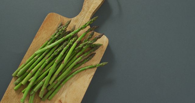 In this image, fresh green asparagus is seen on a wooden chopping board over a grey background. The asparagus is neatly arranged and emits a sense of freshness and healthiness, emphasized by its vibrant green color. The simple but striking contrast between the grey background and the natural texture of the wooden board makes the vegetables stand out. This type of image can be effectively used in culinary blogs, recipe books, healthy eating guides, diet plans, vegan and organic product advertisements, and food preparation tutorials to highlight fresh produce and healthy living.