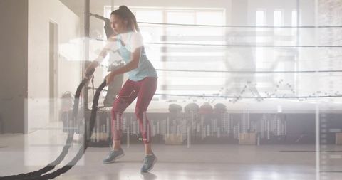 Female athlete swinging battle ropes in sunlit gym for cardio strength training