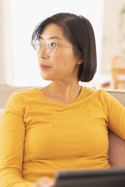 Asian woman sitting on a sofa in a living room, using a tablet and smiling. Ideal for lifestyle, technology, and home comfort themes. Perfect for promoting digital devices, home decor, and leisure activities.