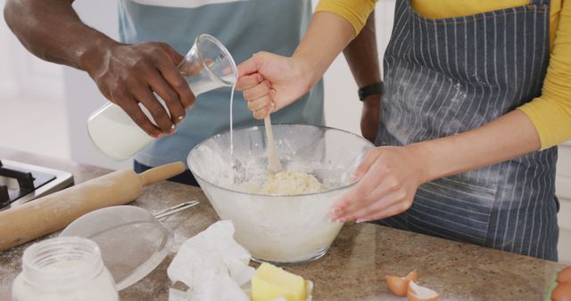 Couple baking together in kitchen while mixing dough in glass bowl. Hands working together showcasing teamwork. Fresh ingredients like flour, eggs, milk on countertop indicates baking process. Suitable for concepts of togetherness, culinary activities, home cooking, and domestic lifestyle.