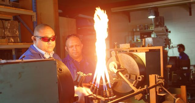 Technicians stand in a workshop, using a gas torch generating a bright, controlled flame. Wearing safety glasses and blue uniforms, they observe the process intently, working on metal machinery. This stock photo can be used for themes related to industrial work environments, safety in technical professions, teamwork in engineering, manufacturing processes, and training materials for metalwork or welding courses.