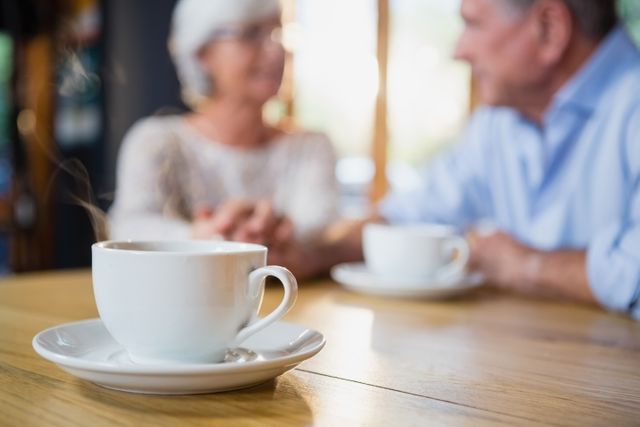 Close-up of a steaming coffee cup on a wooden table in a café, with a senior couple holding hands and conversing in the background. Ideal for use in advertisements for cafés, articles on senior lifestyle and relationships, or promotional materials for coffee brands.