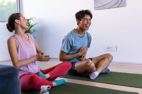 Diverse friends laughing during home yoga session, man clutching stomach, woman clapping