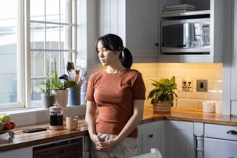 Asian woman leaning on kitchen counter with french press, pensive morning in cozy kitchen