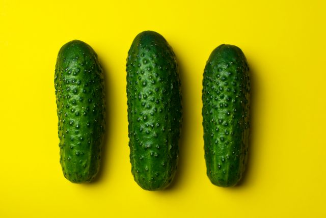 Three fresh cucumbers placed in a row against a bright yellow background. Ideal for use in food photography, health and nutrition articles, summer recipe illustrations, organic produce advertising, and culinary blogs. This vibrant and contrasting color scheme makes the vegetables stand out, highlighting their texture and freshness.