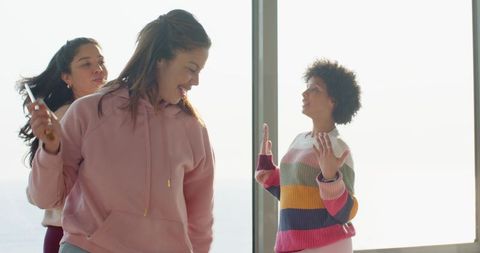 Laughing multiracial women dancing by sunlit windows in pink hoodie and striped sweater