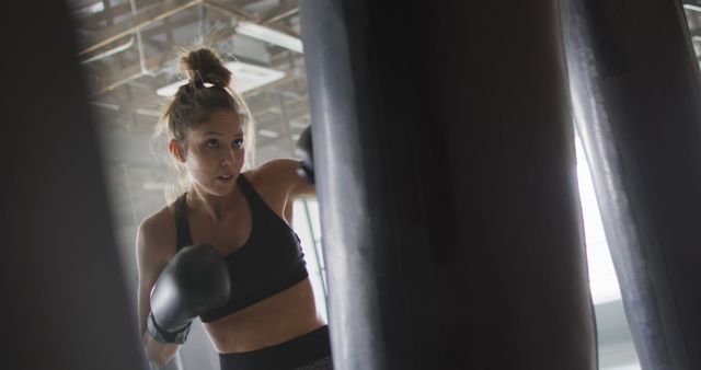 A determined woman wearing boxing gloves punching a boxing bag in a gym environment. Suitable for advertising fitness programs, showcasing gym facilities, promoting women's strength and empowerment, or inspiring people to engage in physical activity and training.